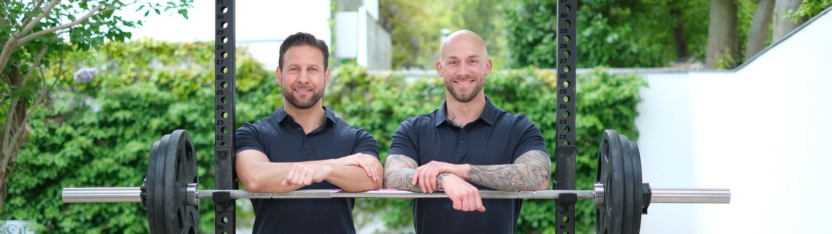 Christian Blisse und Julian Schlüter, Trainerteam, bei einer Hantelstange im Freien.