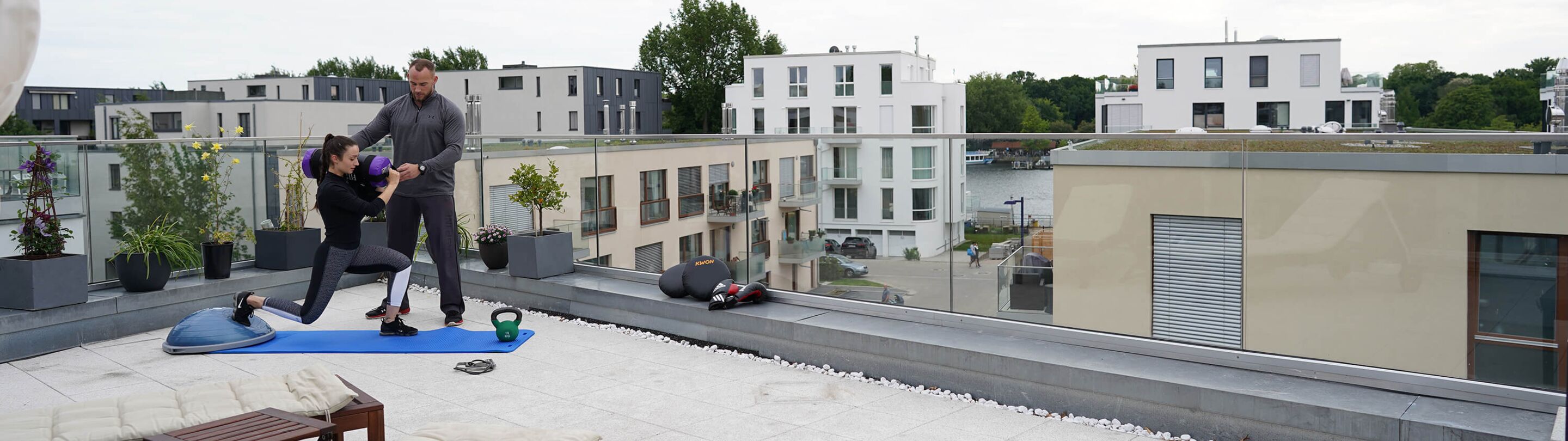 Trainer und Klientin auf Dachterrasse, Klientin auf Fitnessmatte mit Trainings-Utensilien.